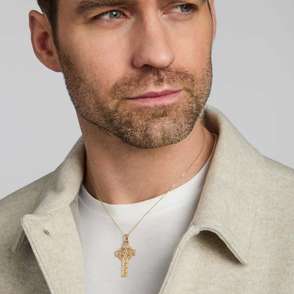A close-up of a man wearing a gold cross pendant necklace over a white t-shirt and a light beige coat, set against a solid light gray background.